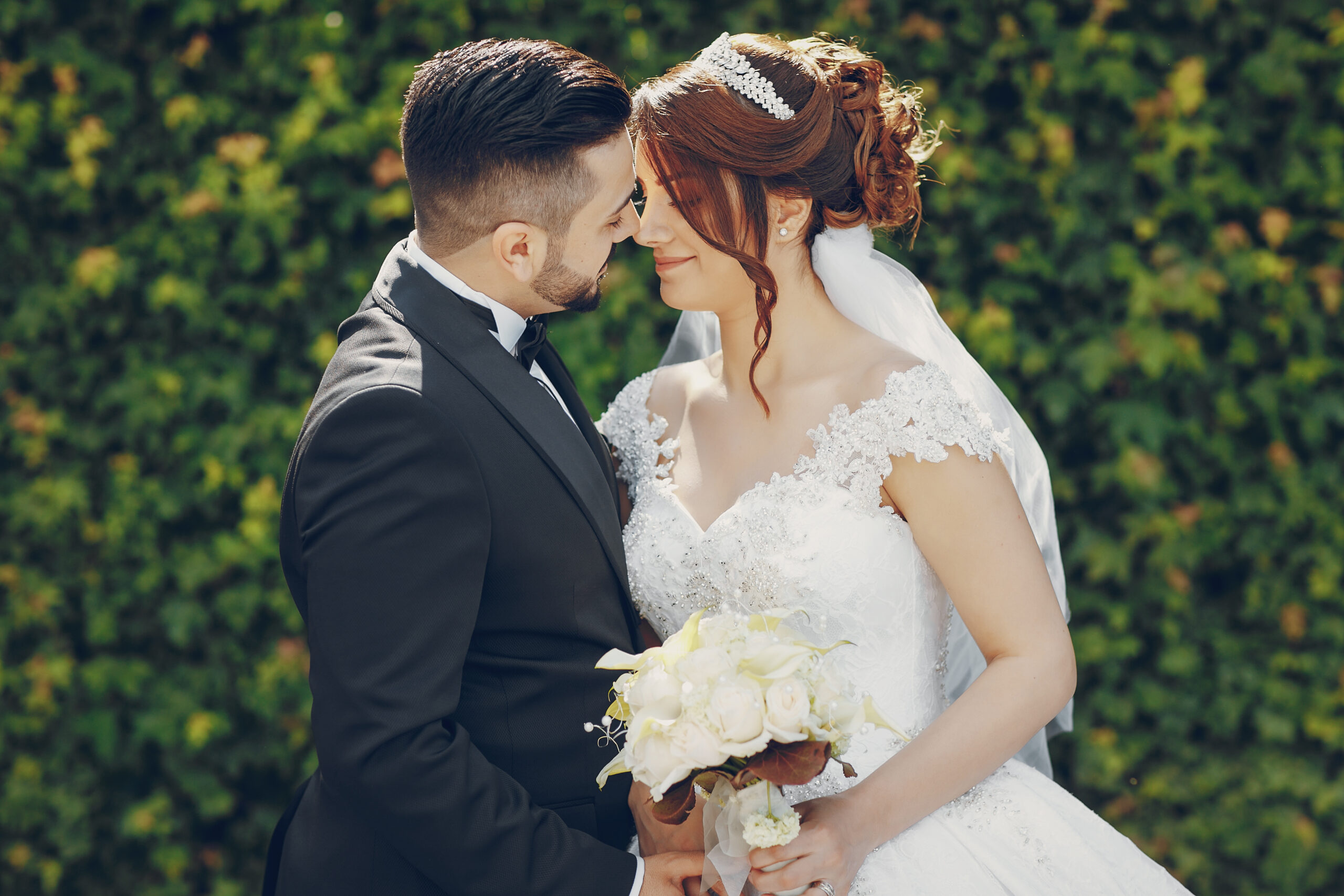 A beautiful young Turkish man wearing a black suit and a beard standing in the park along with his beautiful bride in a white dress and a bouquet of flowers